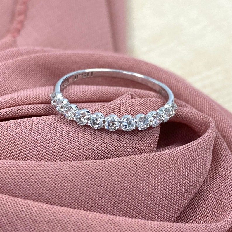 Pu&ograve; includere: Una delicata fascia d'argento con una fila di piccoli diamanti rotondi incastonati in una linea continua. L'anello &egrave; mostrato su uno sfondo di tessuto rosa morbido.