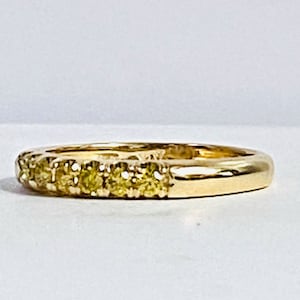 Puede incluir: Un anillo de banda dorada con una hilera de peque&ntilde;as piedras preciosas redondas de tono amarillo engastadas a lo largo de la mitad frontal. El metal tiene un acabado pulido y reflectante, y las piedras est&aacute;n alineadas en una fila recta.