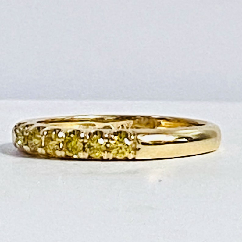 Puede incluir: Un anillo de banda dorada con una hilera de peque&ntilde;as piedras preciosas redondas de tono amarillo engastadas a lo largo de la mitad frontal. El metal tiene un acabado pulido y reflectante, y las piedras est&aacute;n alineadas en una fila recta.