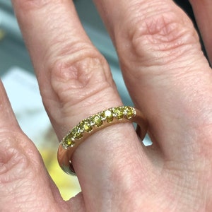 Puede incluir: Un anillo de banda de oro con una hilera de siete peque&ntilde;as piedras preciosas amarillas de corte redondo engastadas a lo largo de la mitad superior de la banda. El metal tiene un acabado pulido y las piedras est&aacute;n dispuestas en un engaste compartido.