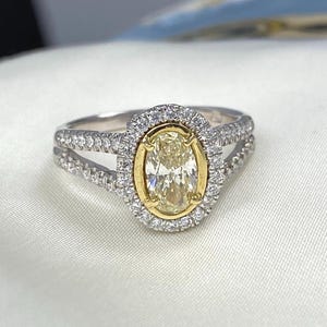 May include: A ring featuring a central oval-cut yellow stone set in a yellow gold bezel, surrounded by a halo of small white stones. The split shank band is crafted from silver-toned metal and is encrusted with small white stones along both sides.