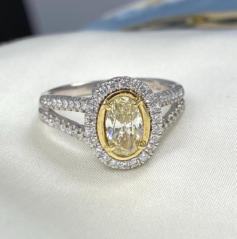 May include: A ring featuring a central oval-cut yellow stone set in a yellow gold bezel, surrounded by a halo of small white stones. The split shank band is crafted from silver-toned metal and is encrusted with small white stones along both sides.