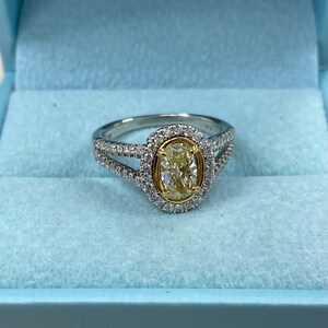 May include: An oval-cut yellow diamond ring set in a yellow gold bezel, surrounded by a halo of small white diamonds. The band is made of silver-toned metal and features a split-shank design encrusted with additional small white diamonds.