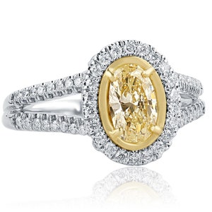May include: An oval-cut yellow diamond ring set in a yellow gold bezel, surrounded by a halo of small round white diamonds. The split shank band is crafted from white metal and is encrusted with rows of small round white diamonds.