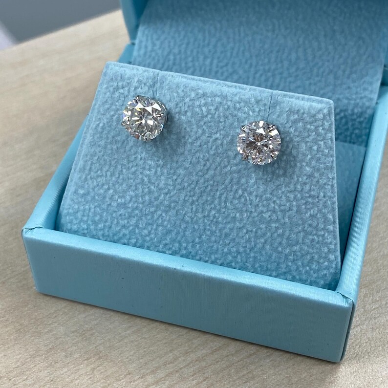 May include: A pair of diamond stud earrings in a light blue velvet box. The earrings are round and have a clear, sparkling appearance.