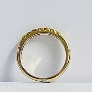 Puede incluir: Un anillo de banda dorada con una hilera de siete peque&ntilde;as piedras preciosas redondas de tono amarillo engastadas a lo largo del arco superior. El metal tiene un acabado pulido y reflectante, manteniendo una forma circular lisa.