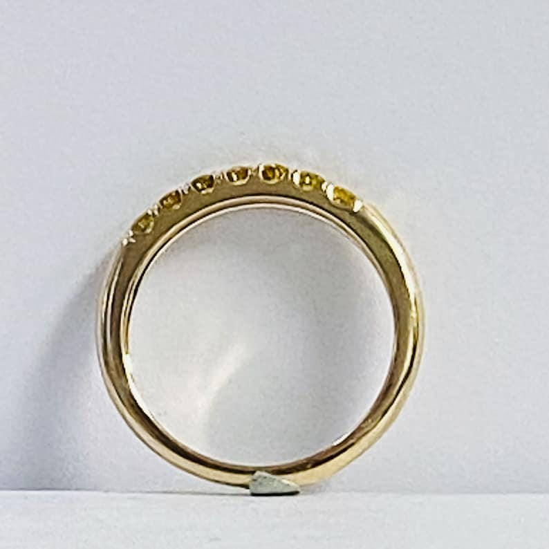 Puede incluir: Un anillo de banda dorada con una hilera de siete peque&ntilde;as piedras preciosas redondas de tono amarillo engastadas a lo largo del arco superior. El metal tiene un acabado pulido y reflectante, manteniendo una forma circular lisa.