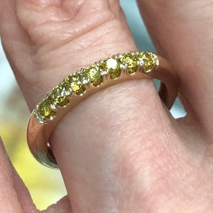 Puede incluir: Un anillo de banda de oro con una fila de siete piedras amarillas de corte redondo en un engaste de garras compartidas. El metal tiene un acabado pulido y las piedras est&aacute;n dispuestas linealmente a lo largo de la mitad superior de la banda, mostrando un tono amarillo vibrante.