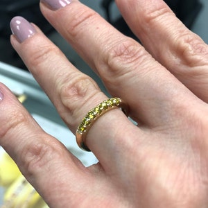 Puede incluir: Un anillo de banda dorada con una hilera de peque&ntilde;as piedras preciosas de color amarillo brillante engastadas a lo largo de la mitad superior de la superficie met&aacute;lica. El anillo tiene un acabado pulido y un perfil redondeado.