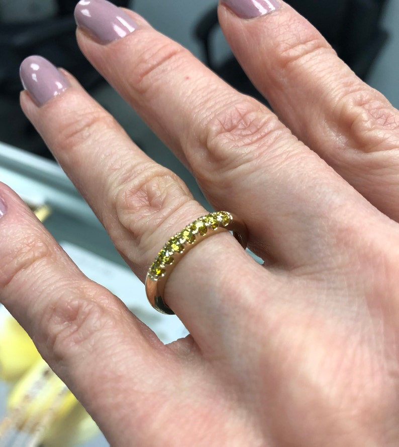 Puede incluir: Un anillo de banda dorada con una hilera de peque&ntilde;as piedras preciosas de color amarillo brillante engastadas a lo largo de la mitad superior de la superficie met&aacute;lica. El anillo tiene un acabado pulido y un perfil redondeado.