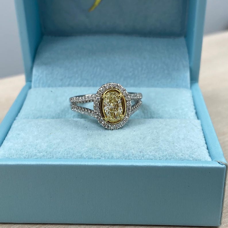 May include: An oval-cut yellow diamond ring set in a gold bezel, surrounded by a halo of small white diamonds. The band is made of silver-toned metal and features a split-shank design encrusted with additional small white diamonds.