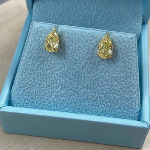 May include: A pair of yellow pear-shaped diamond stud earrings set in gold. The earrings are presented in a light blue jewellery box with a velvet interior.
