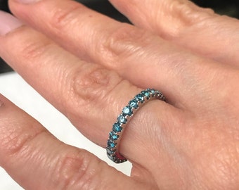 May include: A silver ring with a band of small, round, blue gemstones. The ring is on a person's finger, with the hand in soft focus. The gemstones are a vibrant, eye-catching color.