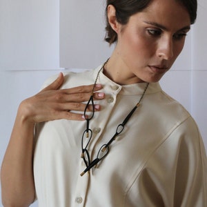 May include: A woman wearing a white collared shirt with a black leather necklace featuring gold accents. The necklace has a geometric design with multiple loops and teardrop shapes.