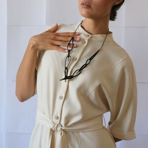 May include: A woman wearing a white dress with a black and gold necklace. The necklace is made of linked oval shapes with gold accents.
