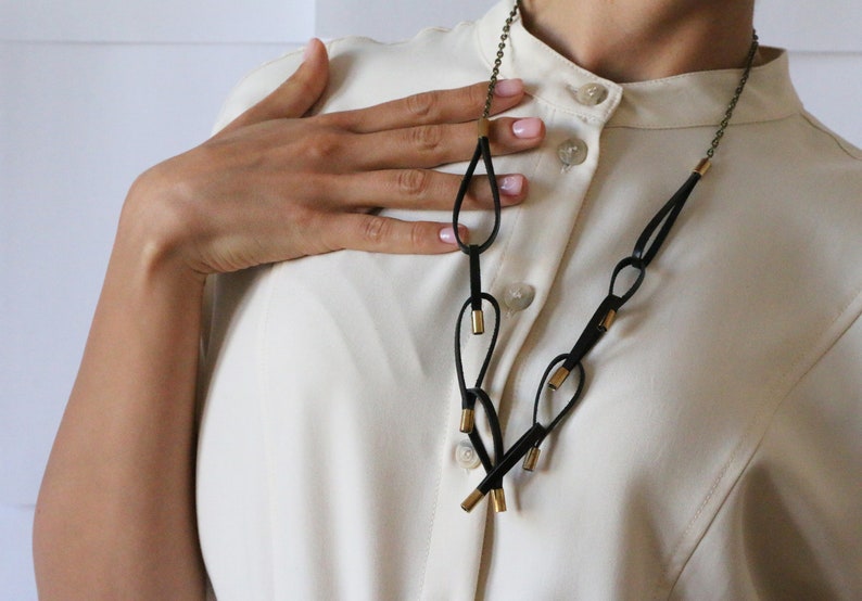 May include: A black leather necklace with gold accents. The necklace features a series of loops and is worn by a person with a white shirt.