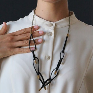 May include: A black leather necklace with gold accents. The necklace features multiple loops and is worn by a person with a white shirt.