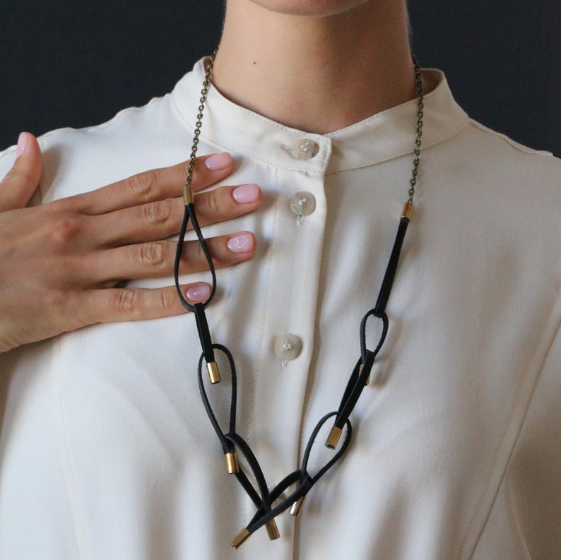 May include: A black leather necklace with gold accents. The necklace features multiple loops and is worn by a person with a white shirt.