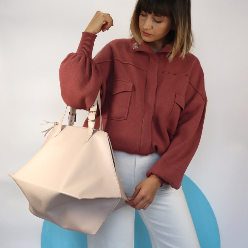May include: A light pink geometric shaped tote bag with a brown leather strap and buckle closures. The bag is being held by a person wearing a red sweater and white pants.