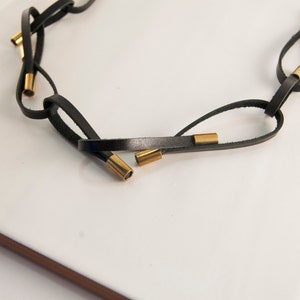 May include: A black leather necklace with gold metal accents. The necklace is made of multiple strands of leather that are interwoven and knotted together. The gold accents are small, cylindrical pieces that are spaced evenly along the necklace.