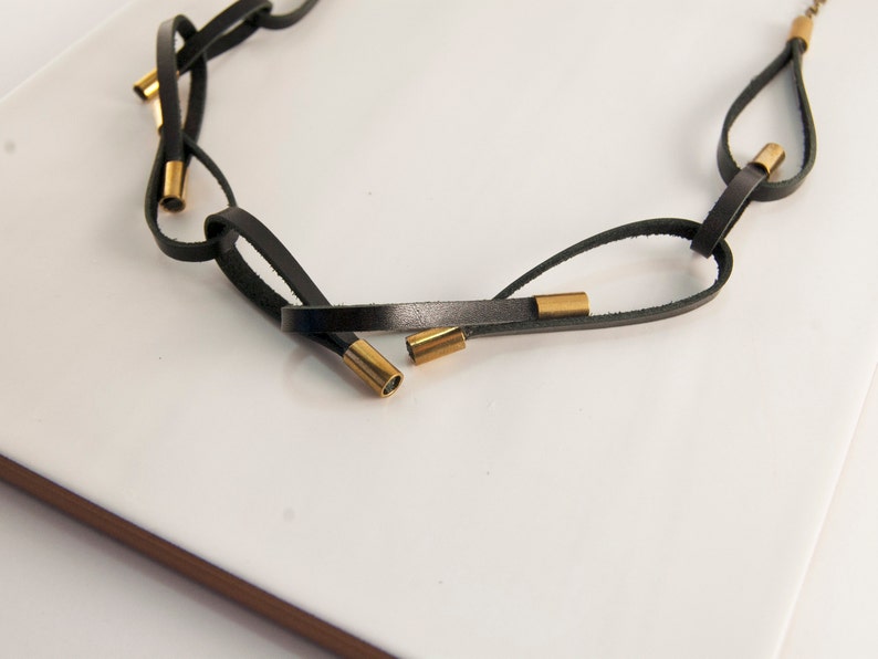 May include: A black leather necklace with gold metal accents. The necklace is made of multiple strands of leather that are interwoven and knotted together. The gold accents are small, cylindrical pieces that are spaced evenly along the necklace.