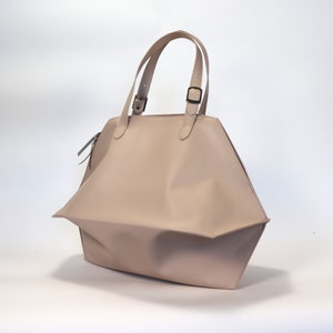 May include: A light beige leather tote bag with two top handles and a buckle closure. The bag has a slightly slouched look and a geometric shape.