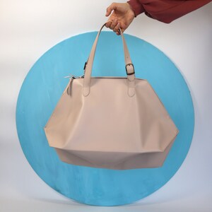 May include: A pale pink leather tote bag with a geometric shape and two top handles. The bag has a zipper closure and a buckle detail.