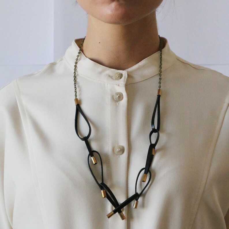 May include: A black leather necklace with gold accents. The necklace features a series of connected loops and is worn by a person with a white shirt.