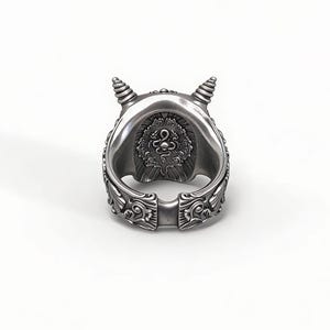 Op de afbeelding: Een zilverkleurige ring met een gedetailleerd ontwerp. De ring heeft een centraal embleem en decoratieve patronen langs de band. Twee kleine, spiraalvormige hoorns bevinden zich aan de bovenkant. De ring staat tegen een witte achtergrond.