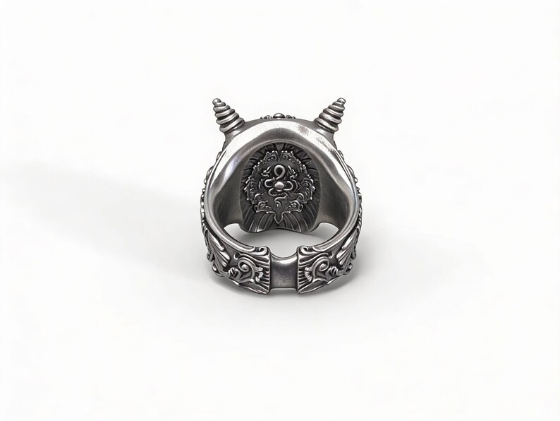 Op de afbeelding: Een zilverkleurige ring met een gedetailleerd ontwerp. De ring heeft een centraal embleem en decoratieve patronen langs de band. Twee kleine, spiraalvormige hoorns bevinden zich aan de bovenkant. De ring staat tegen een witte achtergrond.