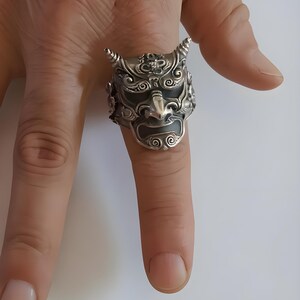 Op de afbeelding: Een gedetailleerde zilveren ring met een Japans demonenmaskerontwerp. De ring heeft ingewikkelde details, waaronder hoorns, een grimas en sierlijke patronen. De ring wordt om een vinger gedragen en toont zijn grootte en vakmanschap.