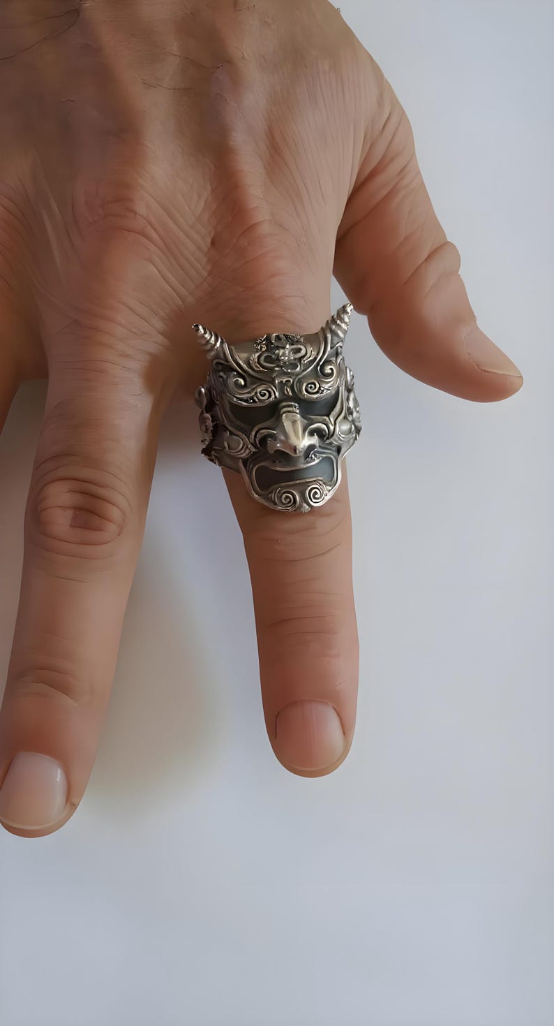 Op de afbeelding: Een gedetailleerde zilveren ring met een Japans demonenmaskerontwerp. De ring heeft ingewikkelde details, waaronder hoorns, een grimas en sierlijke patronen. De ring wordt om een vinger gedragen en toont zijn grootte en vakmanschap.