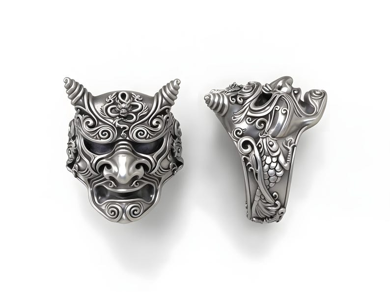 Op de afbeelding: Twee zilveren ringen met ingewikkelde ontwerpen. E&eacute;n ring is een gedetailleerd masker met hoorns, de andere ring heeft een gestileerd gezicht met sierlijke patronen. De ringen staan tegen een witte achtergrond.