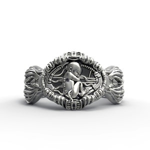 May include: A silver ring with a detailed design featuring a baby figure inside a circular frame. The frame is surrounded by intricate patterns and the ring has a wide band.