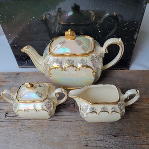 May include: A vintage tea set comprising a teapot, creamer and sugar bowl. The set is made of white porcelain with gold trim and an iridescent glaze. The teapot has a square body and a lid with a gold knob. The creamer and sugar bowl are both round.