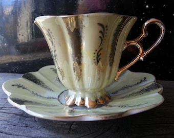 May include: A pale yellow teacup and saucer set with gold trim and floral designs. The cup has a fluted shape and a delicate handle. The saucer has a scalloped edge.