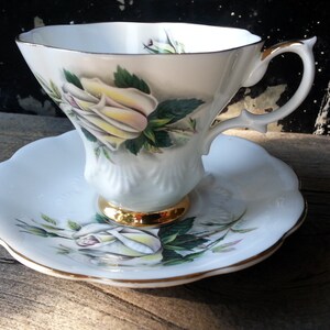 May include: A white teacup and saucer set with a floral rose design. The teacup has a gold-colored base and handle, and the saucer has a scalloped edge. The rose is yellow with green leaves.
