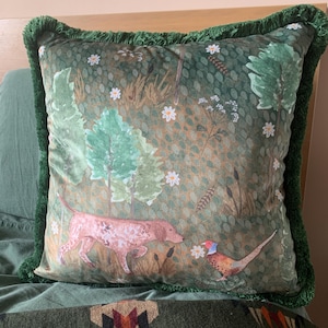May include: Square decorative pillow with a green fringe border. The pillow features a forest scene with a dog, pheasant, trees, and flowers in shades of green, brown, and white. The pillow is placed on a bed.