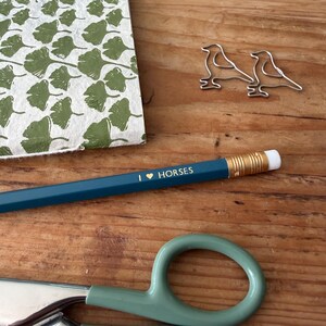 May include: A dark teal pencil with the gold text I HEART HORSES, two silver bird-shaped paper clips, a notebook with a green ginkgo leaf pattern, and a pair of sage green scissors with a large circular handle and a metal blade.