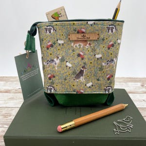 May include: A fabric pencil case with a green zipper and base, featuring a pattern of dogs, sheep, tractors, and badgers on a yellow background. A pencil and a small ruler are visible.
