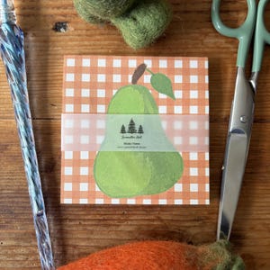 May include: A square pad of sticky notes featuring an orange and white gingham pattern with a large green pear illustration. A translucent paper band wraps around the centre, printed with the text Samantha Hall Sticky Notes and a tree icon.