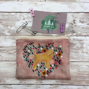 May include: A rectangular zippered pouch made of pink textured fabric featuring a cross-stitched design of a golden dog centred within a heart-shaped wreath of small flowers and green leaves. A card with a tree graphic and the text Samantha Hall.