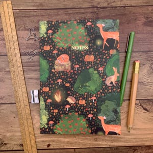 May include: A notebook with a forest design featuring deer, a fox, and a rabbit. The cover has the word "NOTES" and is surrounded by pumpkins, trees, and mushrooms. A green pen and a pencil are next to the notebook.