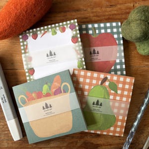 May include: Four square sticky note pads featuring various designs including a green gingham border with fruit and vegetables, a large red apple on a green gingham background, a tan market bag with produce, and a green pear on an orange gingham background.