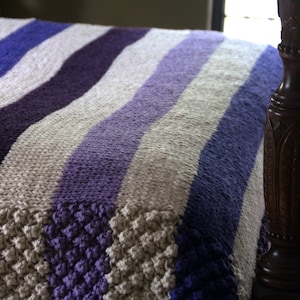 May include: Close-up view of a knitted throw blanket featuring wavy stripes in shades of purple, lavender, and beige. The blanket shows a textured, possibly crocheted, pattern at the bottom. The throw is draped over a dark brown wooden bed frame, suggesting a bedroom setting. The color palette is soft and calming, with a gradient effect in the stripes.