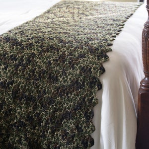 May include: A green, brown, and black crocheted bed runner with a scalloped edge. The runner is on a white bed with a wooden headboard.