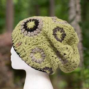 May include: A handmade, slouchy crochet hat in shades of green, brown, and tan. The hat features a floral pattern and a small wooden tag. The brim is black. The hat is displayed on a white mannequin head.