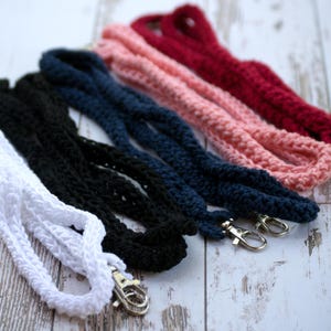 May include: A selection of handmade crochet lanyards in white, black, navy, pink, and burgundy. Each lanyard has a silver-coloured metal clasp for attaching keys or an ID badge. The lanyards are displayed on a light-coloured wooden surface.