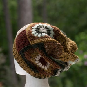 May include: A crochet beanie constructed from multiple square motifs in earthy tones including dark brown, mustard yellow, olive green, and cream. The hat features a textured, multi-colored floral pattern throughout, with a ribbed edge at the base.