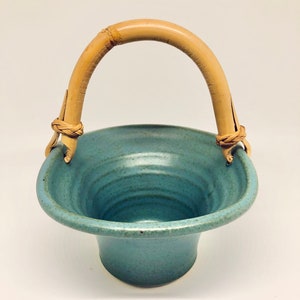 May include: A handmade, teal-colored ceramic basket with a natural bamboo handle. The basket has a flared base and a textured surface, with the handle attached by woven twine. The design is simple and rustic.